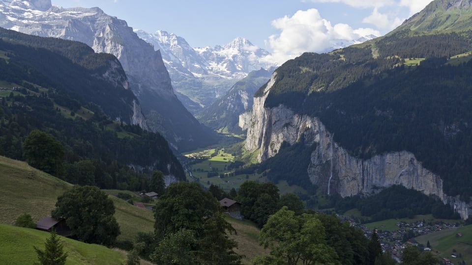 Panoramablick auf ein Bergtal mit Wiesen, Felsen und einem Dorf in den Alpen.