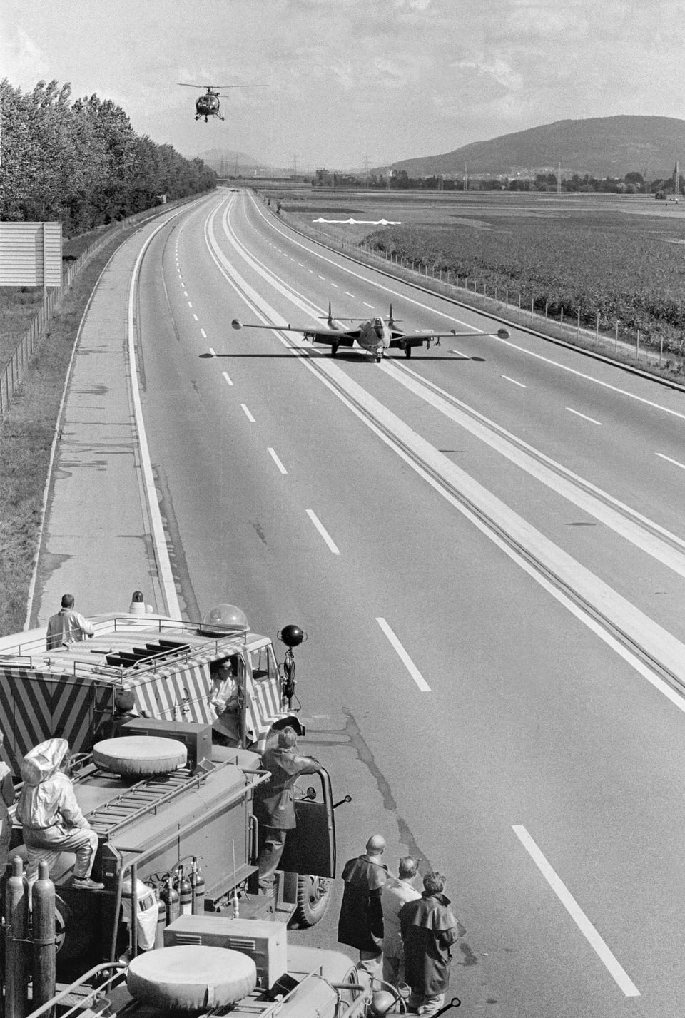 Militärjet rollt auf einer Autobahnstrecke; am Strassenrand sind Fahrzeuge platziert; darüber zirkelt ein Helikopter