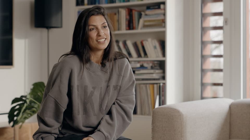 Olivia El Sayed mit langen dunklen Haaren und einem grauen Pullover vor einem Bücherregal sitzend.