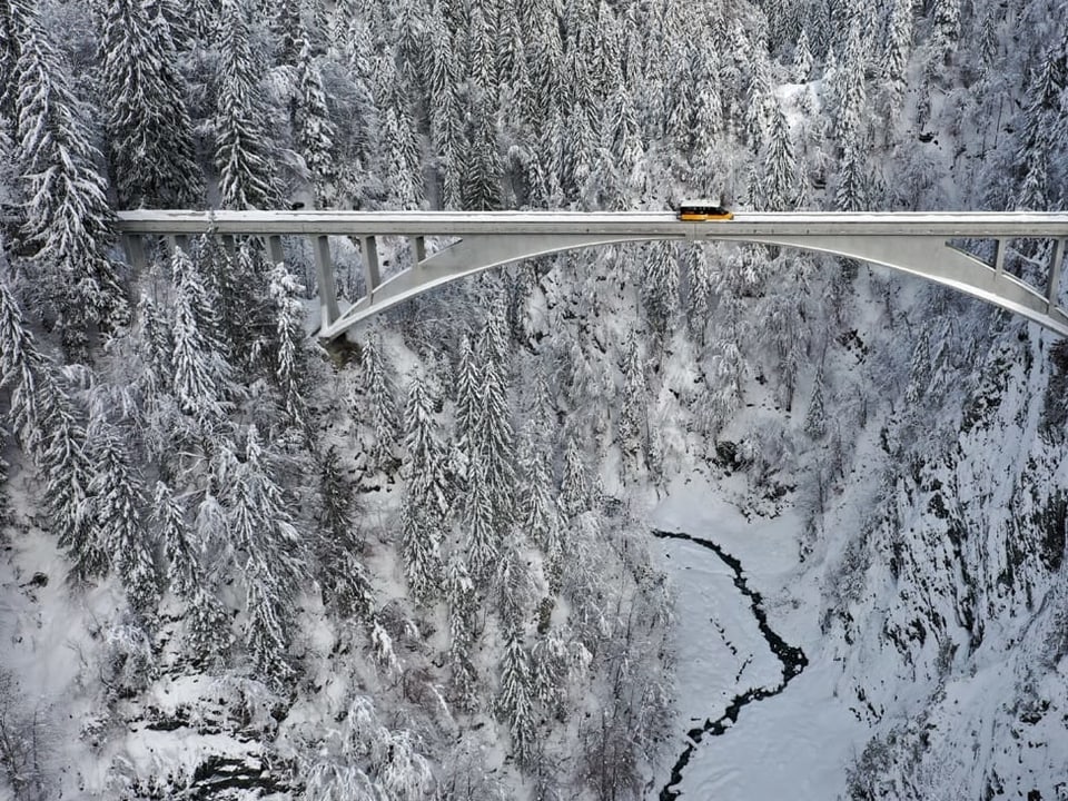 Brücke in verschneiter Landschaft mit Tannen, auf der Brücke ein Postauto