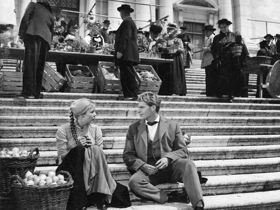 Eine junge Frau und ein junger Mann sitzen auf einer breiten Steintreppe. Neben der Frau zwei grosse Körbe mit Äpfeln. Hinter ihnen steht ein Marktstand.