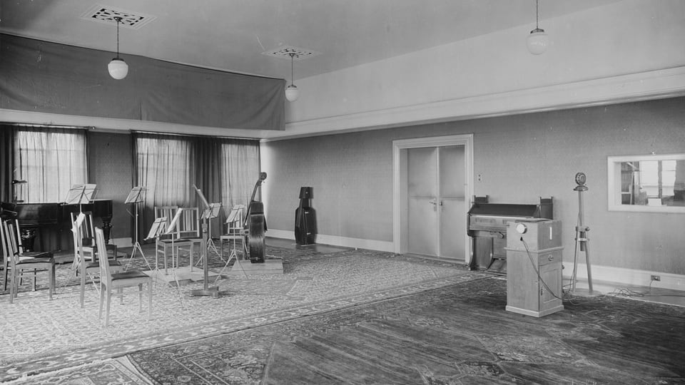 Ein Zimmer mit Stühlen und Notenständern links und einem Klavier und Mikrofon rechts.