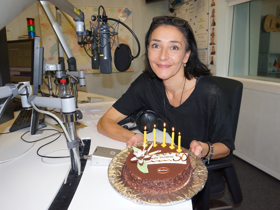 Eine dunkelhaarige Frau mit einer Geburtstagstorte in einem Radiostudio.