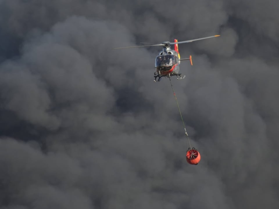 Helikopter trägt Wasserbehälter in dichter Rauchwolke