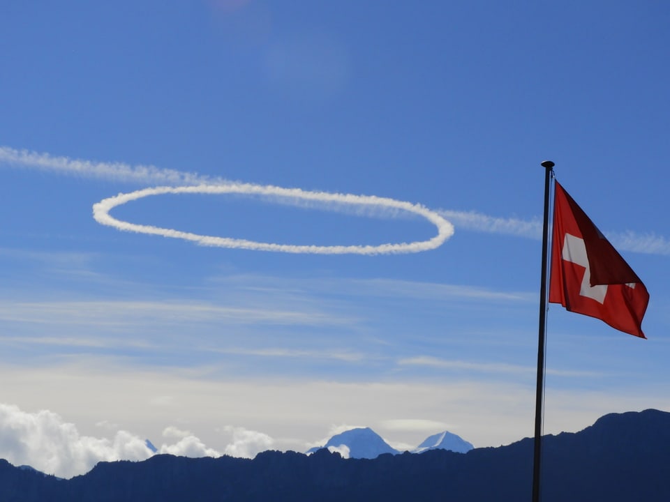 Eine kreisrunde Wolke am sonst blauen Himmel. Im Vordergrund weht die Schweizer Flagge im Wind.