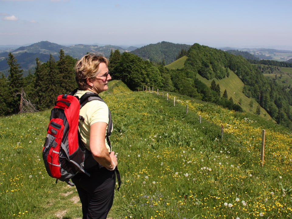 Ruth Breitenmoser mit rotem Rucksack steht auf einem Wanderweg und blickt auf die hügelige Landschaft.