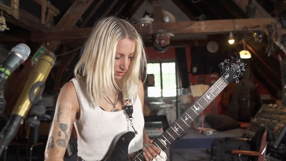 Frau mit Tattoos auf dem Arm und langen Haaren hat eine elektrische Gitarre in der Hand.