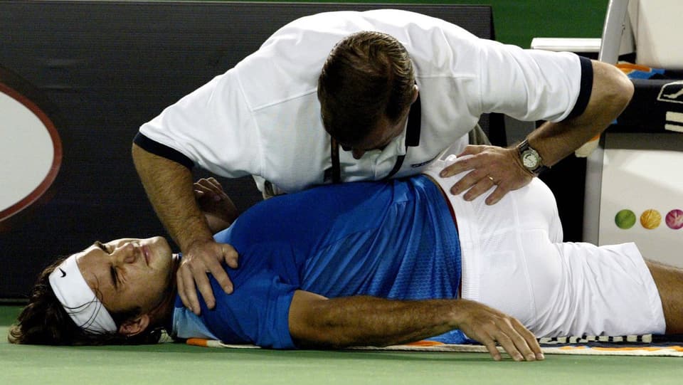 Federer liegt auf dem Boden, ein Physiotherapeut dehnt seinen Rücken, indem er die Hüfte um 90 Grad abdreht.