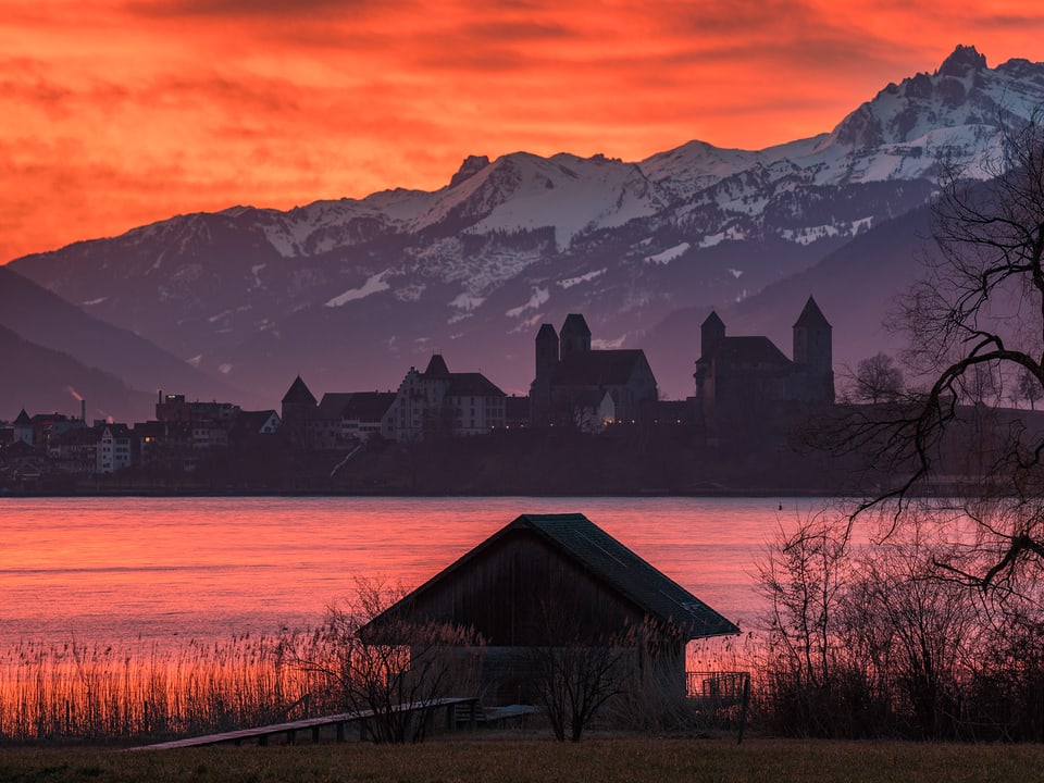 See im Vordergrund, Städtchen im Hintergrund mit Schneebergen. Der Himmel brennt rot und rot spiegelt auch der See. 