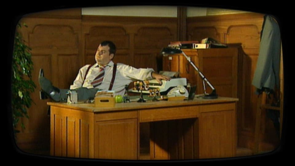 Schauspieler Mike Müller als Beamter, sitzend hinter einem Pult