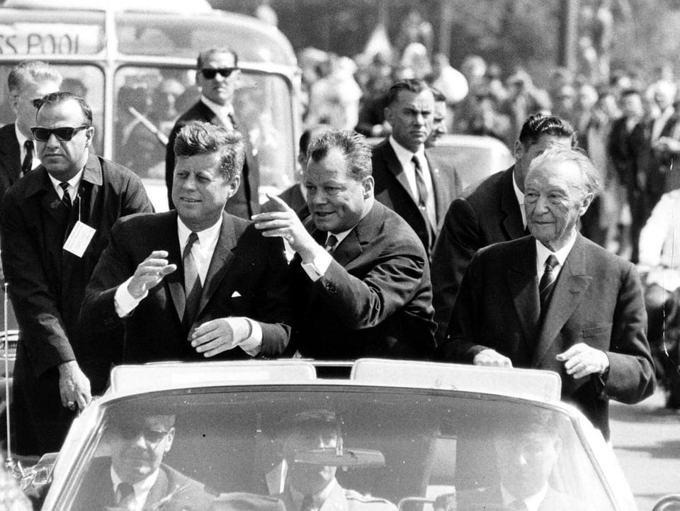 Berlins Bürgermeister Willy Brandt (mitte), der amerikanische Präsident J.F. Kennedy (links) und Bundeskanzler Konrad Adenauer (rechts) fahren in einem Cabriolet durch Berlin. (keystone)