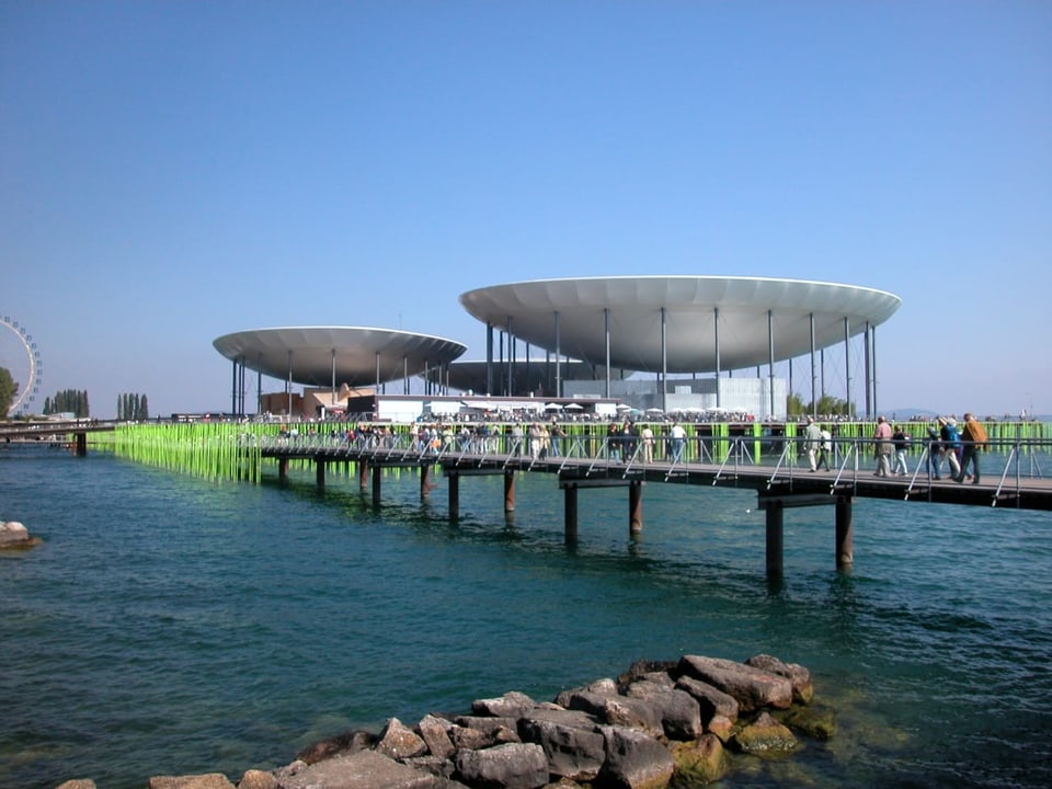 Ein Steg führt über den See zur Arteplage mit der schalenförmigen Überdachung der Arteplage.