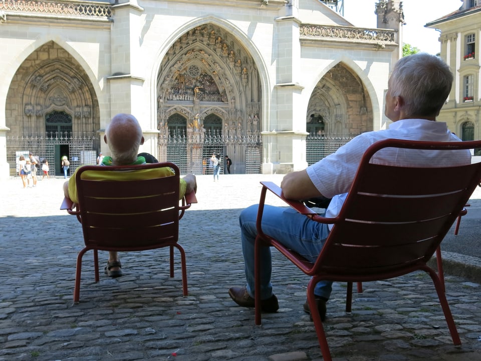 Zwei Herren sitzen auf Stühlen im Schatten und schauen das Münster an.