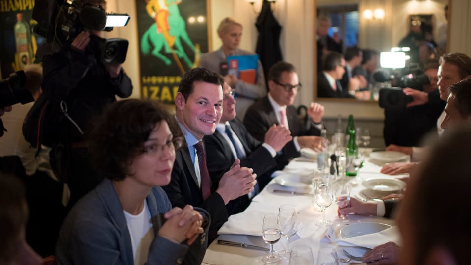 Maudet und seine Frau sitzen in einem Restaurant an einer gedeckten Tafel - im Hintergrund ein Kamermann und eine Frau mit Mikrofon.