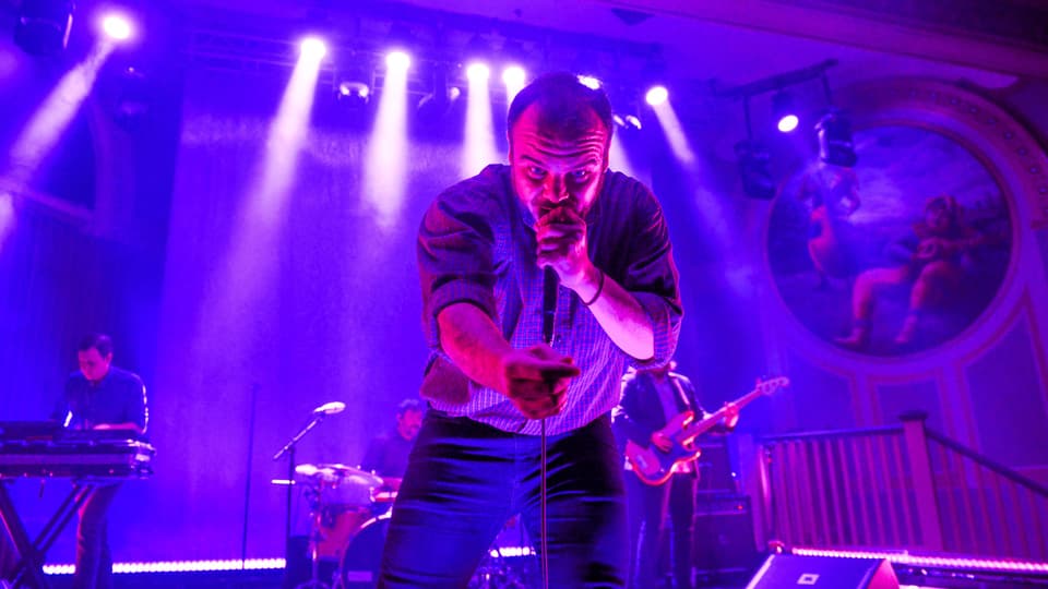 Ein Mann steht auf einer violett beleuchteten Bühne und singt enthusiastisch in ein Mikrofon.