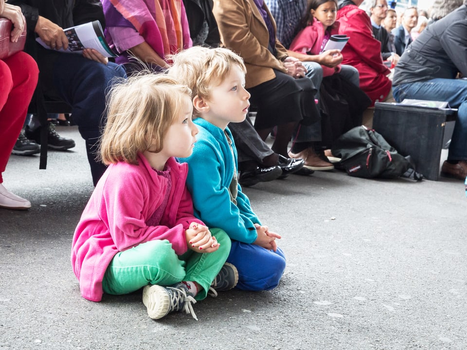 Zwei Kinder sitzen vor der Bühne auf dem Boden und lauschen einem Konzert.