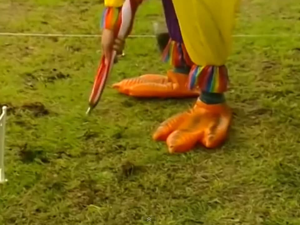 Mann mit Hühnerfuss-Schuhen und Regenschirm versucht, Regenwürmer aus der Erde zu klopfen