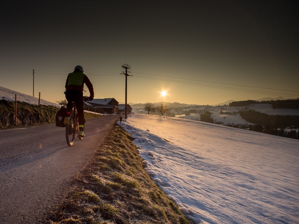 Links Velofahrer auf Strasse. Rechts schneebedecktes Feld. Dahinter Sonne knapp über dem Horizont.
