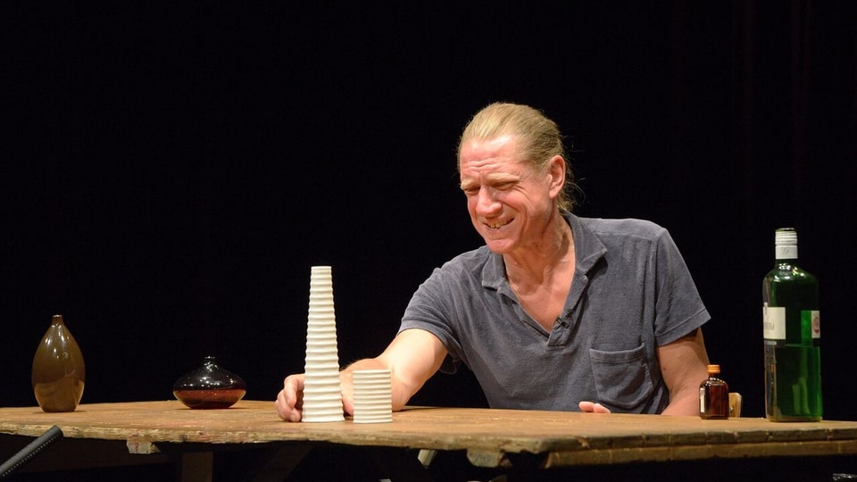 Ein Mann sitzt an einem Tisch mit verzerrtem Gesicht. Auf dem Tisch steht eine Flasche und viele kleine Sachen.
