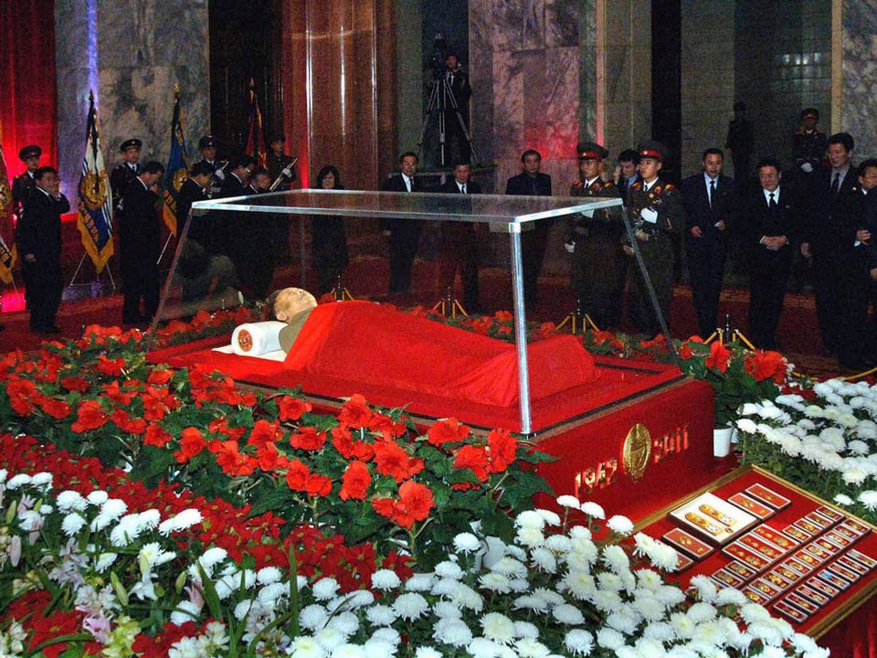 Kim Jong Il in einem Glassarg, darum herum rote und weisse Blumen.