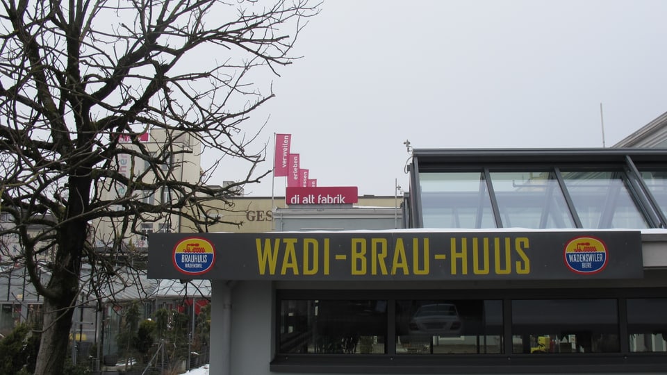 Das Schild des Wädi-Brau-Huus, dahinter ein Schild «di alt fabrik», links ein Baum mit dürren Ästen.