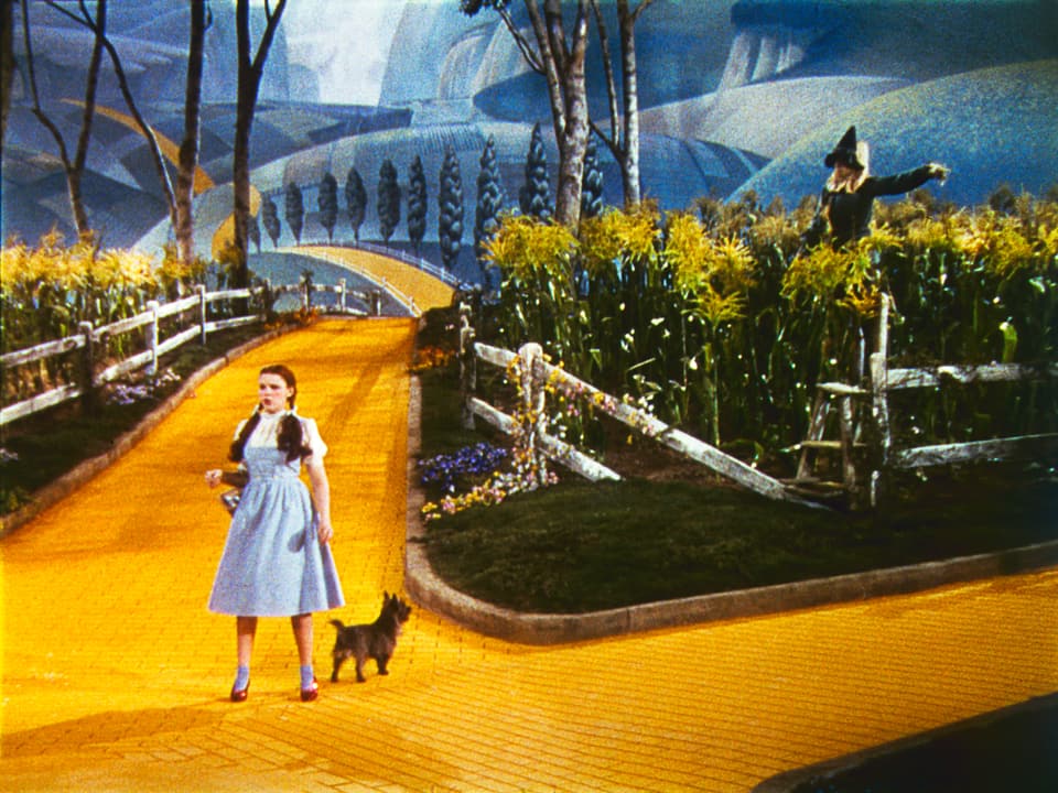 Judy Garland, Toto, Ray Bolger in einer Szene auf einem leuchtend gelben Weg.