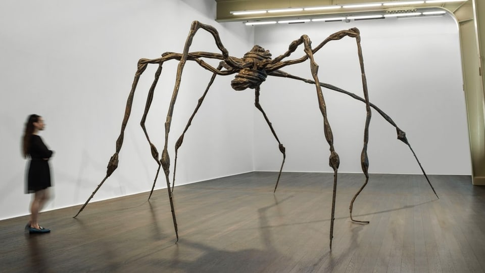 Eine übergrosse Spinne aus Bronze steht in einem Raum neben einer Betrachterin.