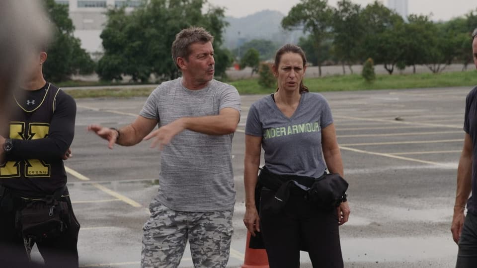 rechts ältere Frau mit zusammengebundenem Haar im T-Shirt, links Mann, Hände in der Luft. Auf einem Parkplatz.