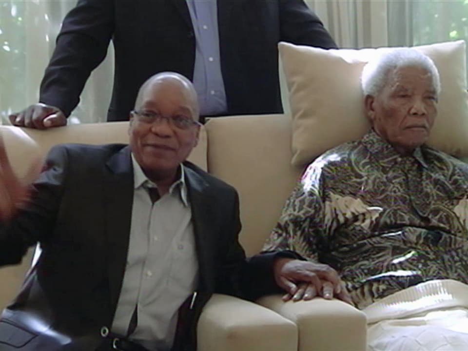Zuma hält Mandelas Hand und winkt in die Kamera. Mandela wirkt apatisch, ein Kissen stützt seinen Kopf