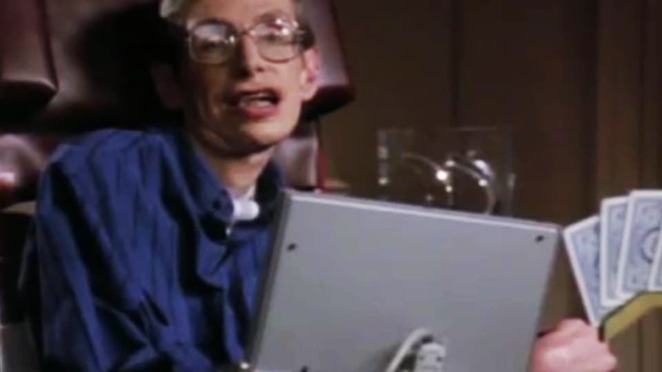 Hawking sitzt hinter seinem Sprachcomputer, Spielkarten in der Hand.