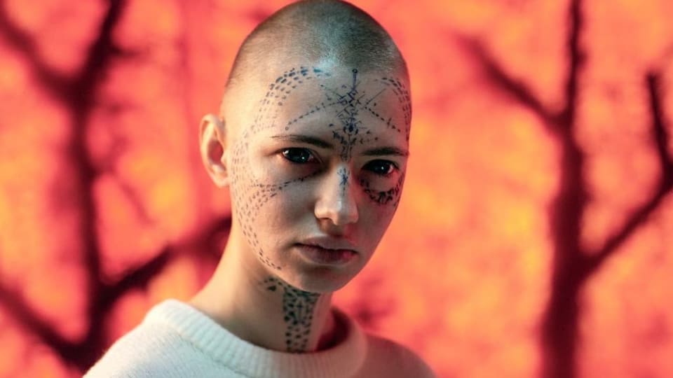 Eine Frau mit kurgeschorenen haaren, Tattoos im Gesicht und schwarzen Augäpfeln.