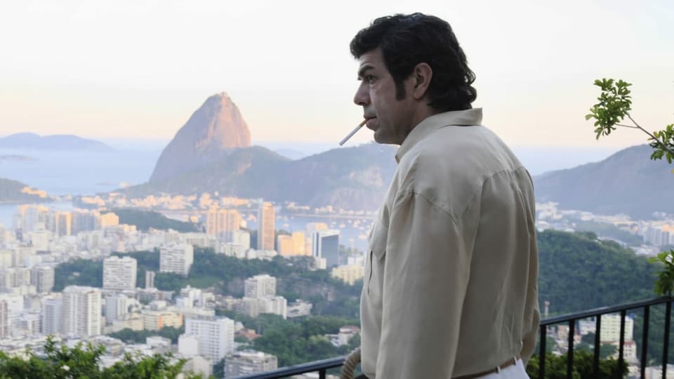 Ein Mann raucht eine Zigarette, im Hintergrund erkennt man Rio.
