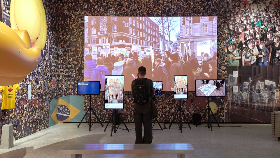 Raum in einem Museum. An der Wand ein grosses Bild einer Menschenmenge, auf verschiedenen Bildschirmen sind Demonstrationen zu sehen. Ein einzelner Mann steht im Raum und schaut auf die Bildschirme.