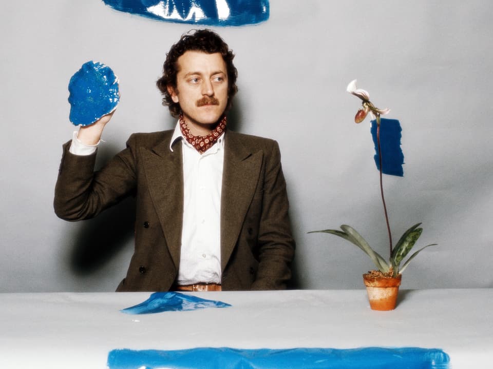 Dieter Meier sitzt an einem Tisch, auf der eine Pflanze steht. Auf dem Tisch und an der Wand sind längliche Farbflecken zu erkennen, in seiner Hand hält er ebenfalls einen "Farbklecks".