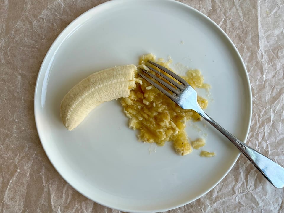 Ein Stück Banane und mit einer Gabel zerdrücktes Bananenmus auf einem weissen Teller.
