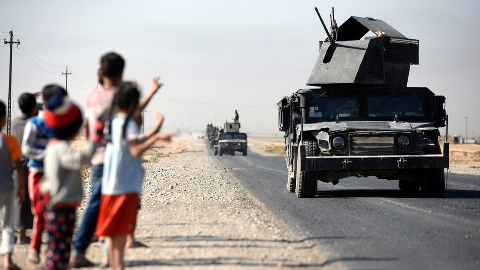 Eine Kolonne gepanzerter Fahrzeuge, am Strassenrand winken Kinder den Soldaten zu.