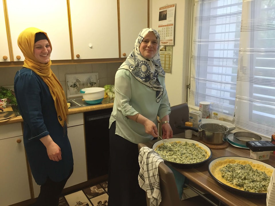 Zwei Frauen mit Kopftuch stehen in einer Küche und befüllen ein mit Teig ausgelegtes Blech mit Lauch und Spinat.
