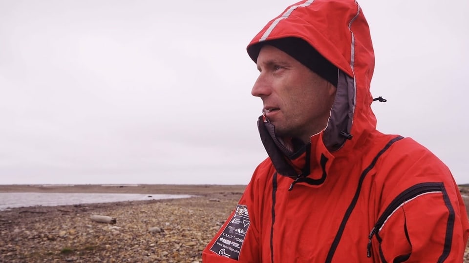 Ein Mann steht an einem steinigen Strand, er hat eine rote Wetterschutzjacke an.