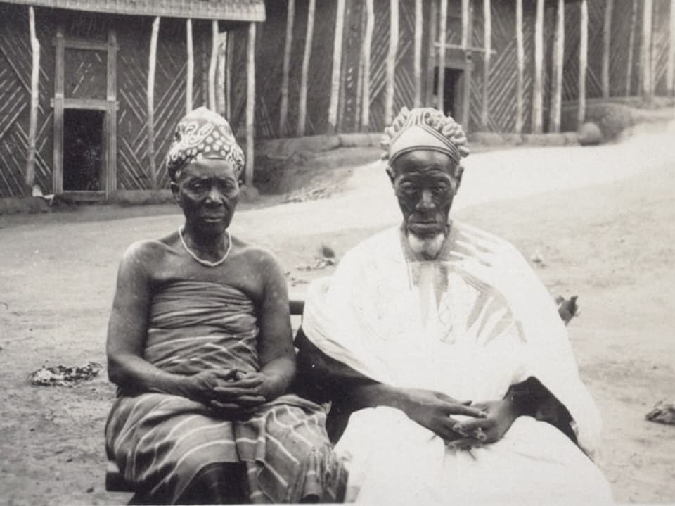 Schwarz-Weiss-Foto eines afrikanischen älteren Paares. Sie sitzen nebeneinander. Beide schauen ernst. 
