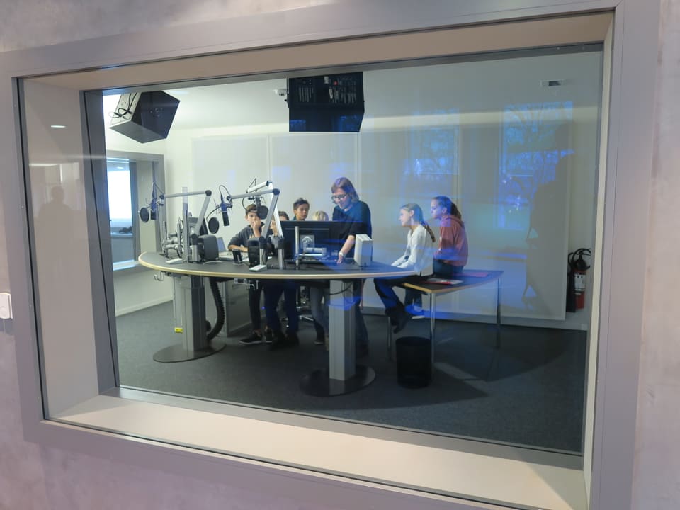 Kinder und ein Redaktor sind durch eine Glasscheibe im Radiostudio zu sehen. 