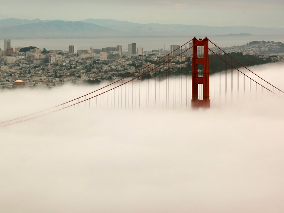 Im Vordergrund dichter Nebel, daraus ragt die rote Golden Gate Bridge hervor. Im Hintergrund die Häuser von San Francisco. 