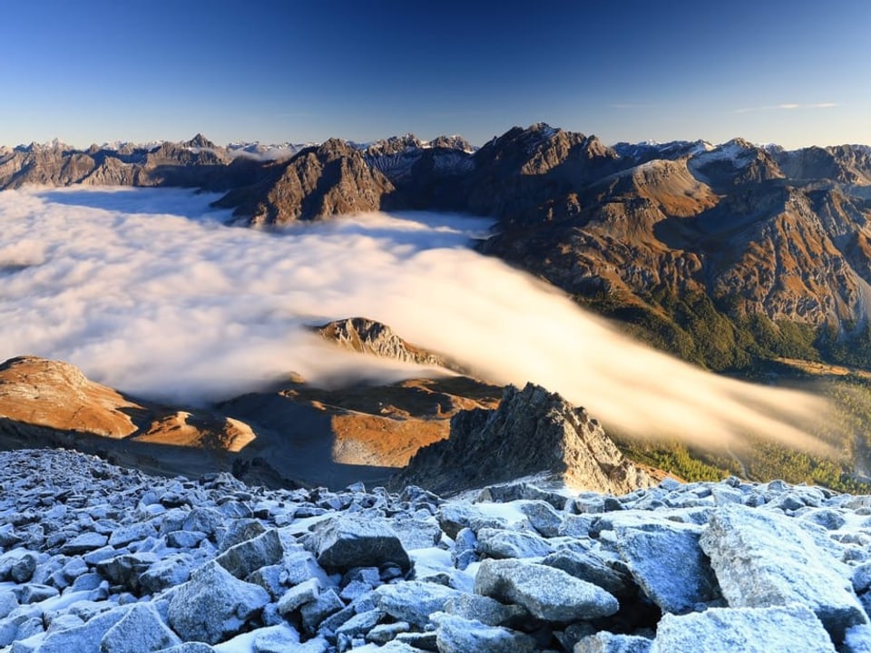 Alpenlandschaft mit Felsen und Nebel im Tal. Darüber wird es bereits sonnig mit blauem Himmel.