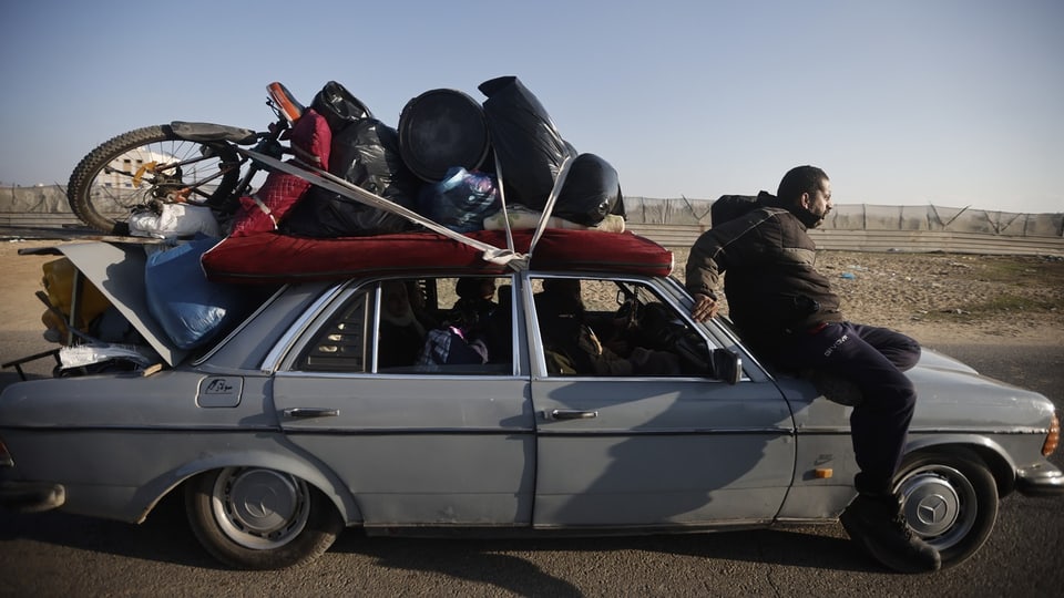 Ein Palästinenser sitzt auf der fronthaube seins Autos. Das Auto ist vollgepackt mit Matratzen, Taschen und einem Velo.