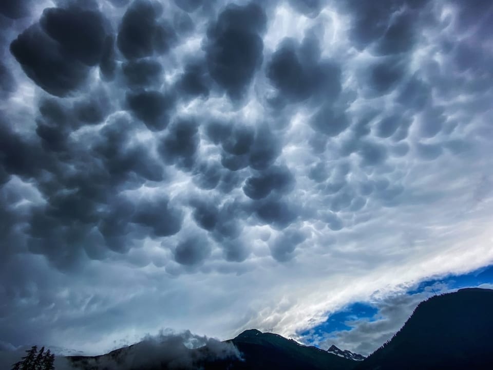 Beutelförmige dunkle Wolken am Himmel, hinten ein Streifen blau, unten Alpen.