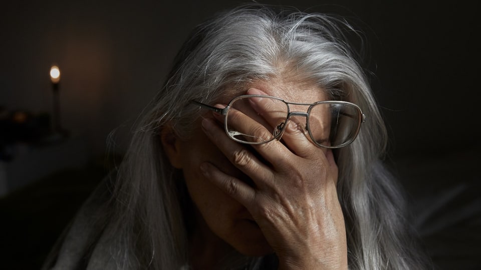 Eine ältere Frau mit langem grauen Haar und Brille hält die Hand vor das Gesicht.