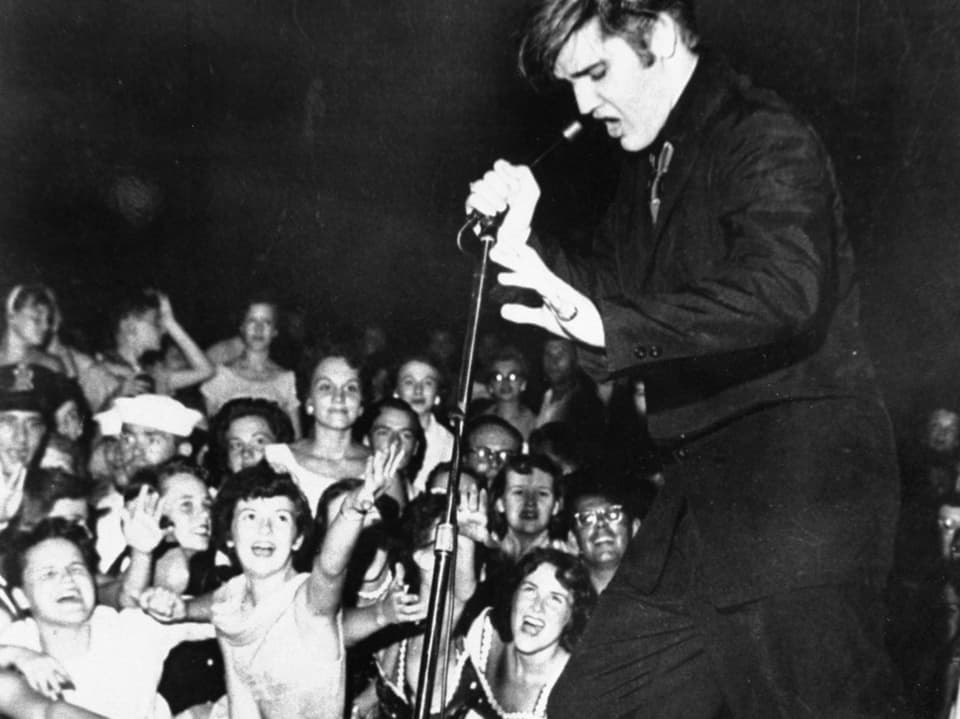 Elvis Presley tanzt auf der Bühne, davor stehen kreischende Fans