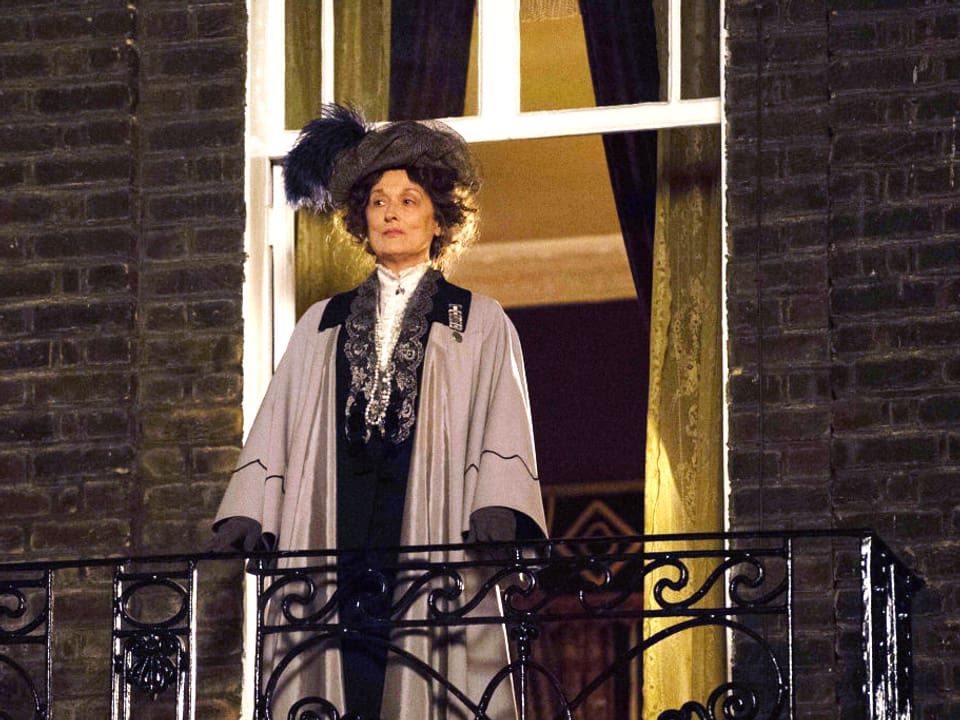 Eine Frau mit langem Gewand und Hut steht auf einem Balkon.