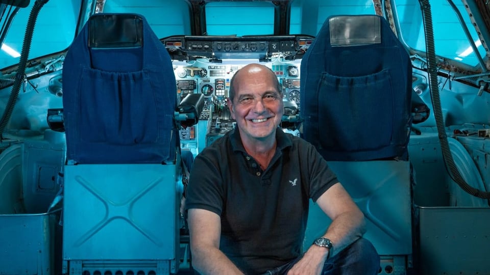 Mann in schwarzem T-Shirt mit Flugzeugmodell vor einem Cockpit.