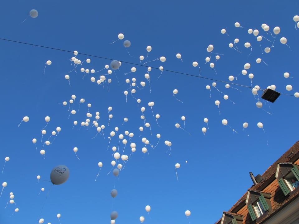Mehrere Dutzend weisse Ballone steigen in den Himmel auf, am rechten Bildrand sieht man ein Hausdach. Ansonsten ist nur der stahlblaue Himmel mit den Ballonen zu sehen.