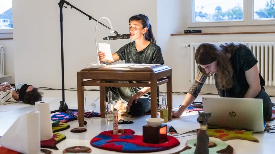 eine junge Frau liest am Mikrofon etwas vor, ein junger Mann werkelt neben an am Laptop